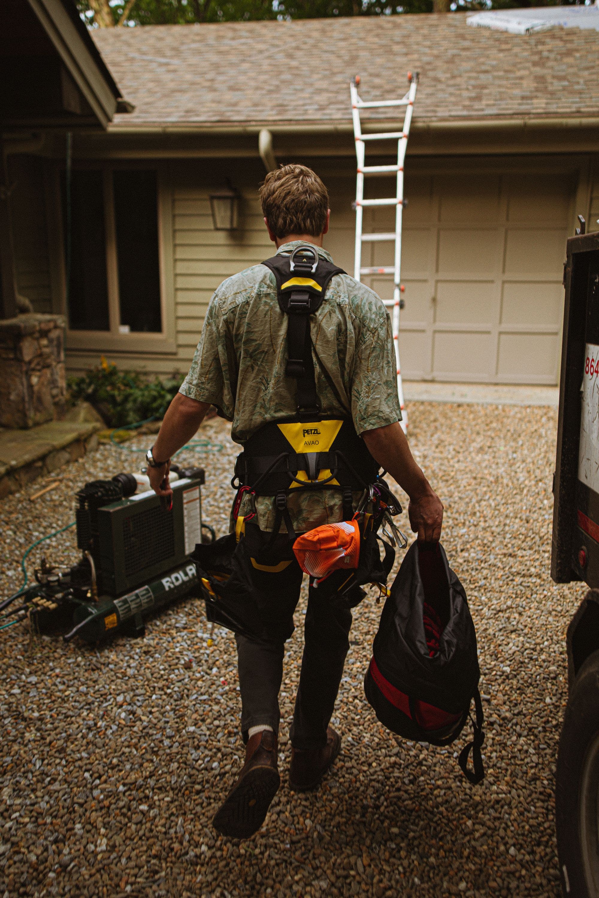 A roofer in a safety harness carrying tools toward a house with a ladder against it